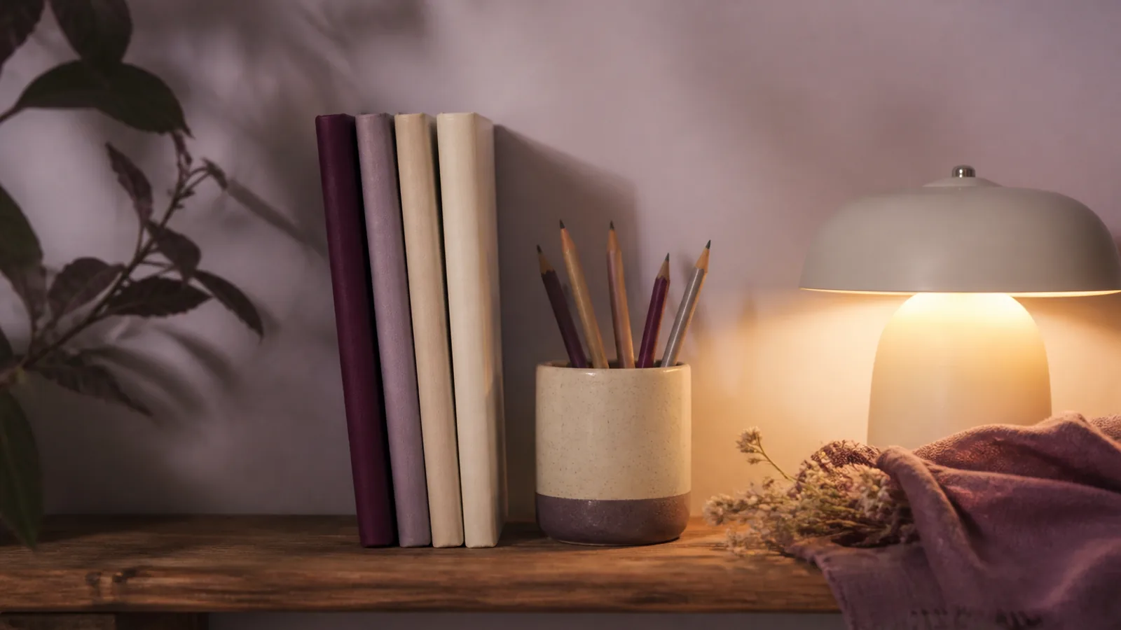 Wooden shelf with four thin travel notebooks, a pencil cup, and a small lamp casting warm light on spines labeled with dusk and dinner notes, soft shadow on a pale wall, 1600x900