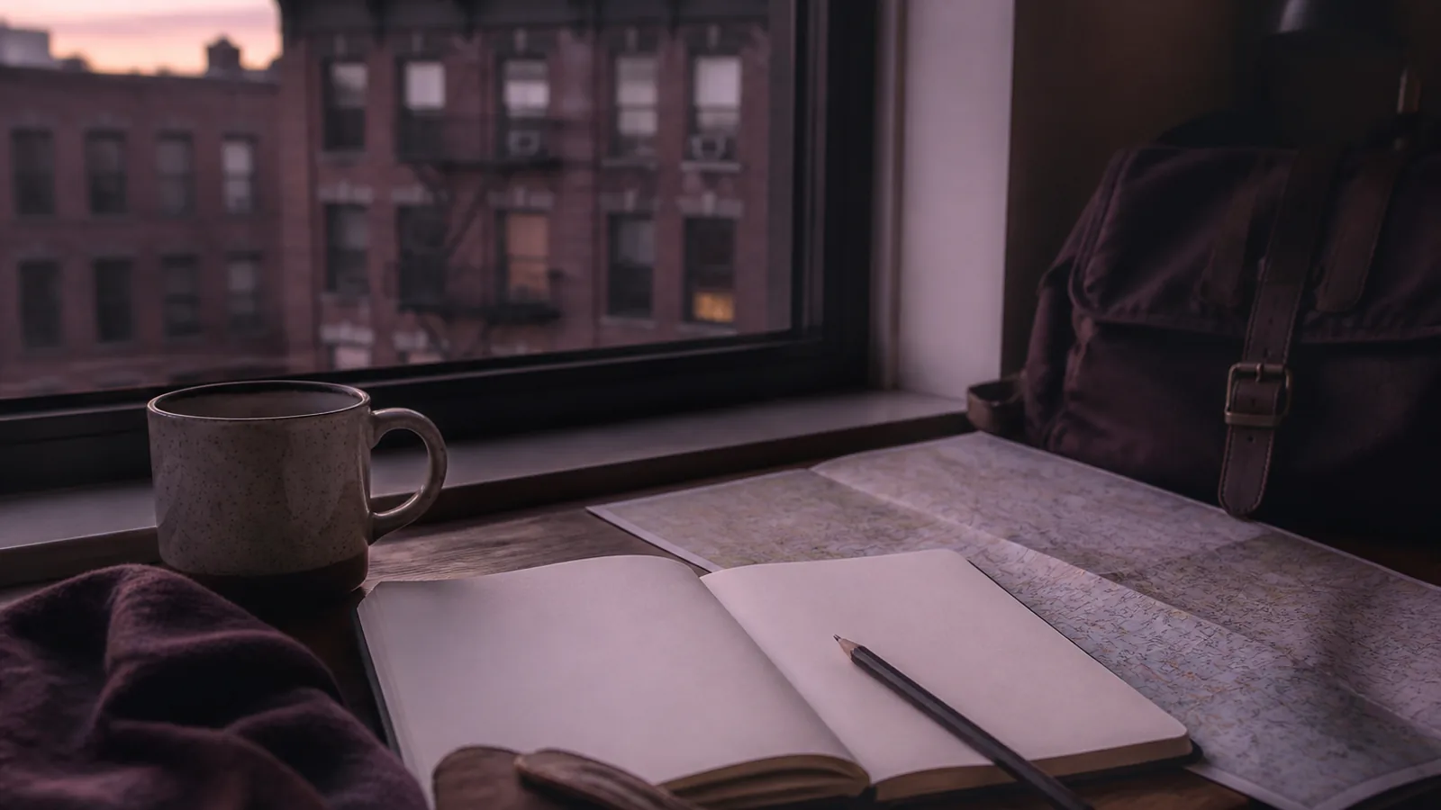 Travel desk with an open notebook, pencil, folded paper map, and a mug beside a window showing early dusk light on brick across the street, 1600x900